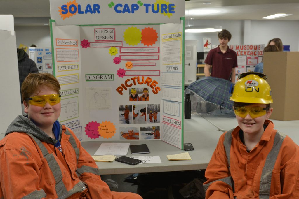 Two male students are dressed in mining gear as they show off their project on solar capture. 