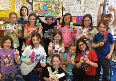 Grade 2 French Immersion students at St. James are gathered together in their classroom smiling while holding up Earth day bookmarks and stickers.