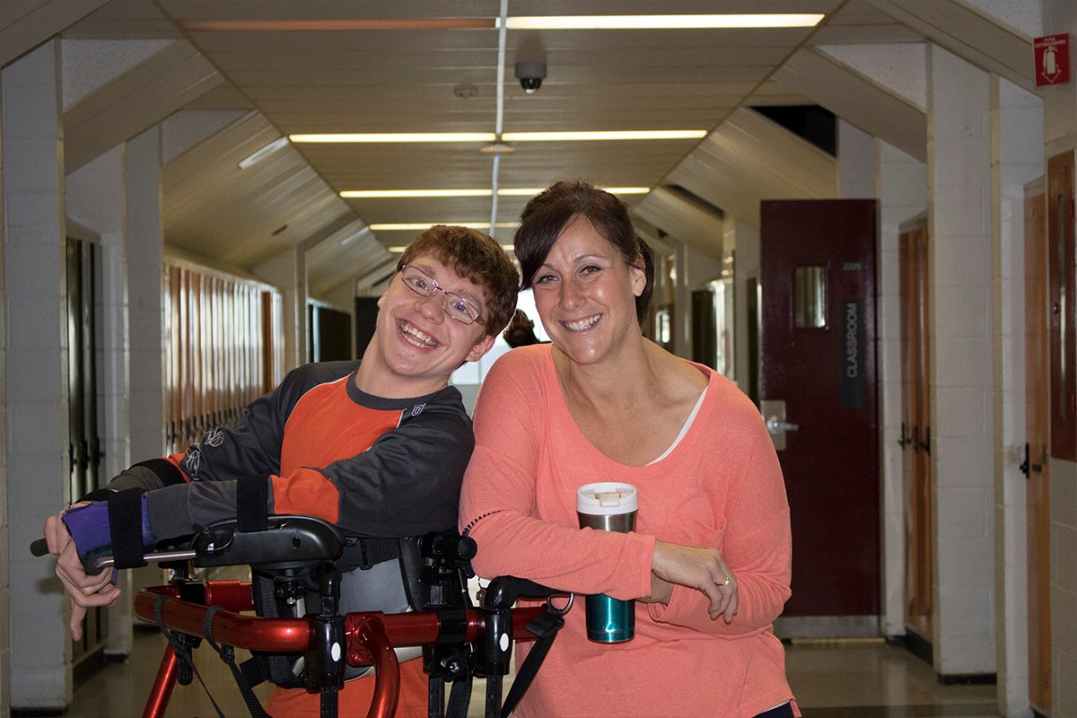 An educational assistant stands smiling with her student in a school hallway.
