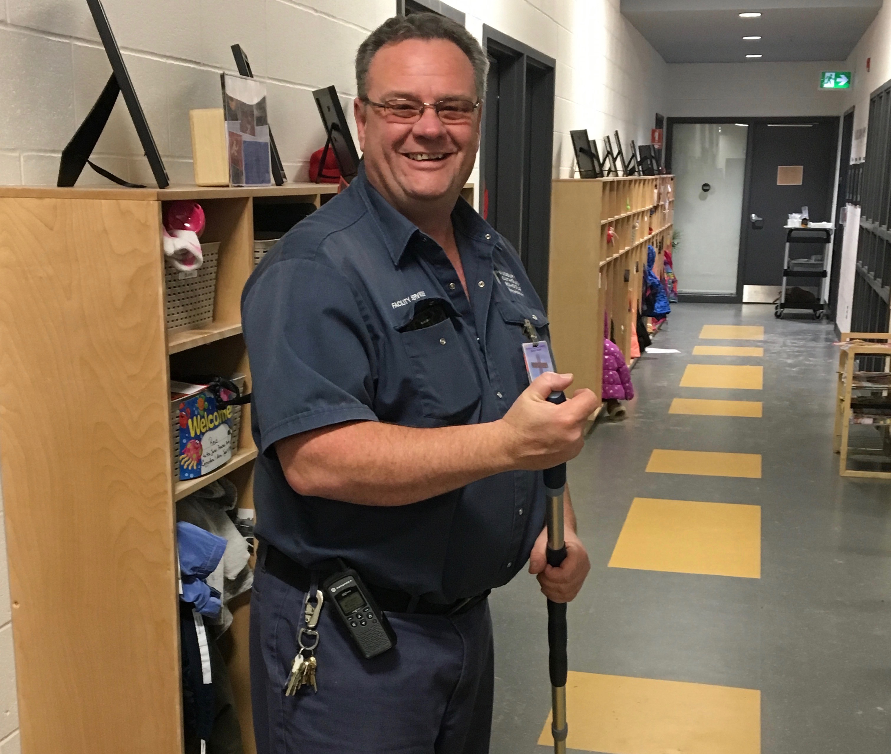 A custodian stands with a large broom smiling.