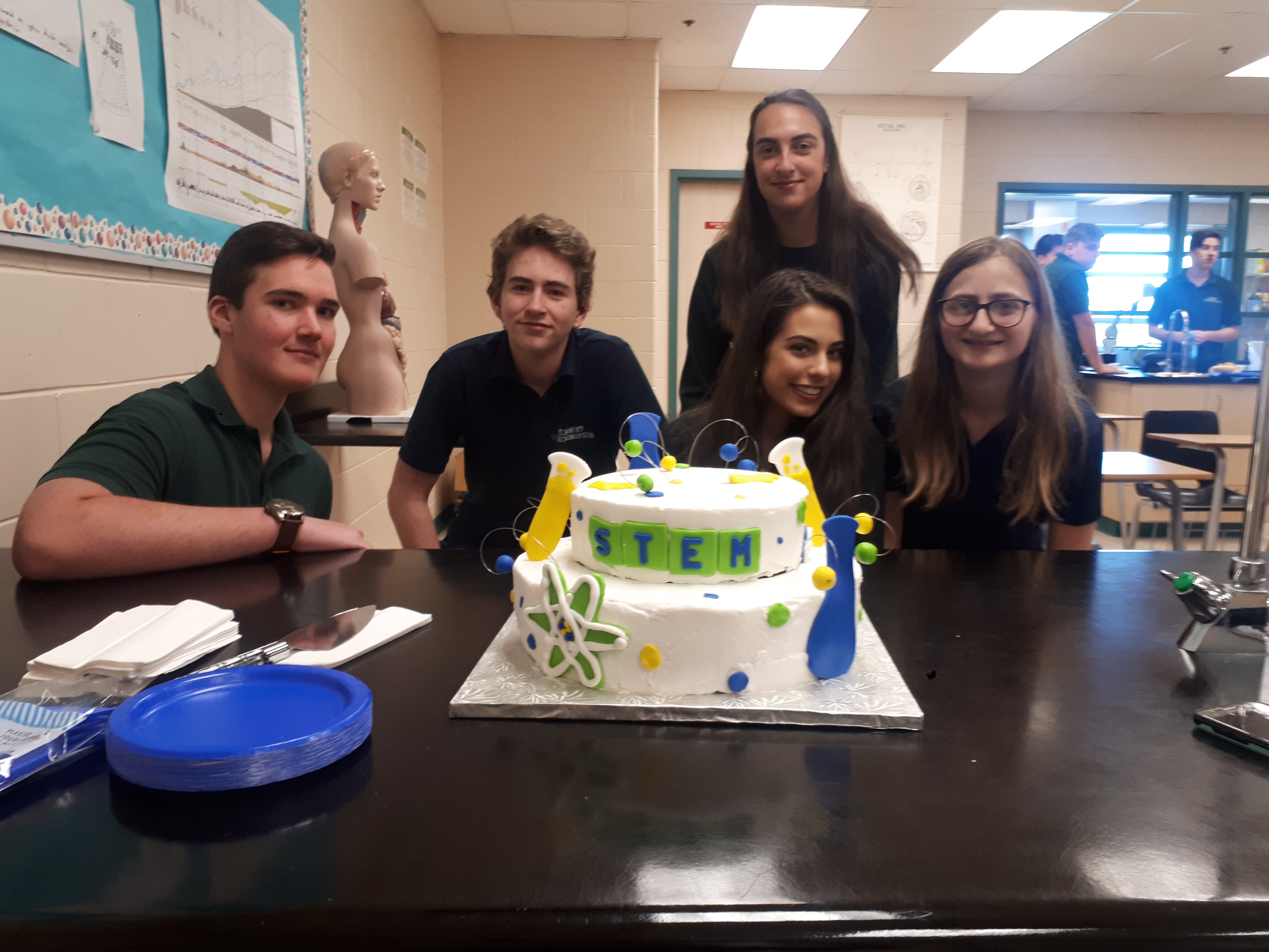 Noah Forteski, Ethan Hodge, Adriana Cimino and Livea Donato with Erika Poirier in the background pose with the STEM Club cake in foreground